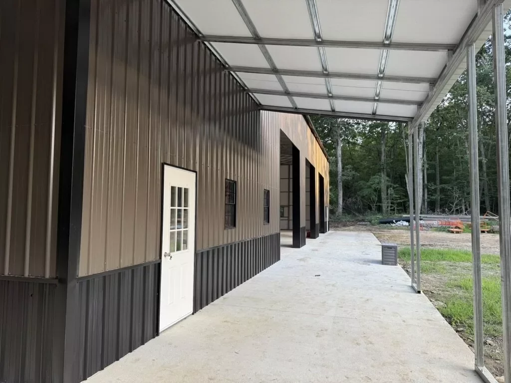 Covered side walkway of the finished metal building with concrete path, white entry door, and dark steel siding